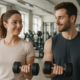 Two friends in a gym lifting dumbbells while smiling at each other.