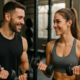 Two fit people in a gym smiling at each other while lifting dumbbells shoulder‑level in a training session of conversation.