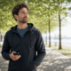 Man in a dark jacket holding a smartphone while strolling along a tree-lined lakeside path, looking ahead thoughtfully.
