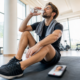 Athlete sitting on a gym mat, drinking water from a bottle with a towel around his neck and a smartphone on the floor nearby.
